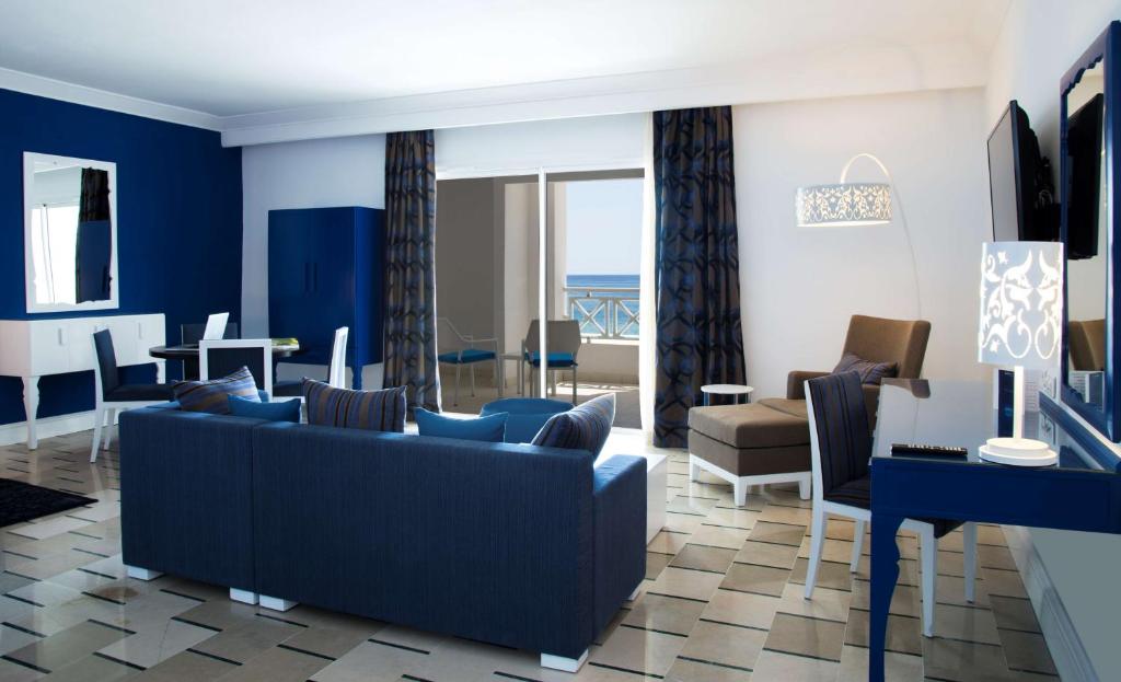 Mūsdienīgs viesistabas interjers ar zilu krāsu paleti, kas ietver komfortablu dīvānu, ēdamgaldu un skatu uz jūru. Gaismas elementi un eleganti akcenti piešķir telpai stilīgu atmosfēru.