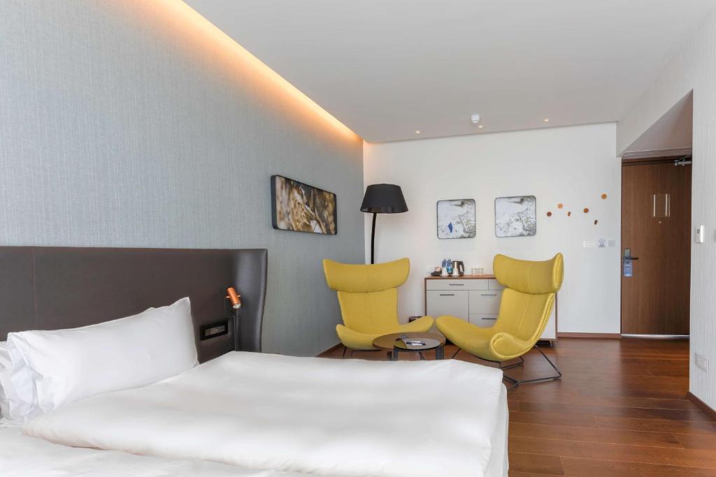 Moderns viesnīcas numurs ar ērtu gultu, divām dzeltenām atpūtas krēslām un elegantu interjeru. Sienas rotā mākslas darbi, radot patīkamu atmosfēru.