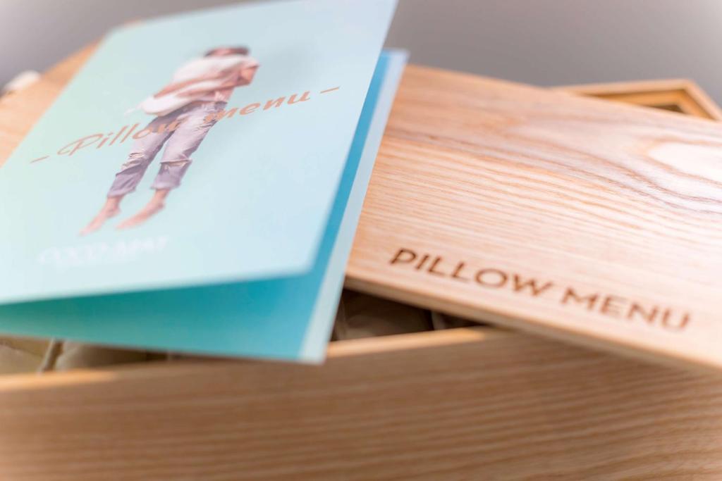 Koka kaste ar izsistiem vārdiem "Pillow Menu" un atvērtu spilvenu izvēles bukletu. Buklets ir gaiši zils ar attēlu, kas atspoguļo komfortu un relaksāciju.