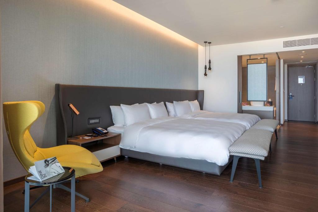 Mūsdienīga viesnīcas numura iekārtojums ar divguļamo gultu, dzeltenu krēslu un koka grīdu. Interjers ir gaišs un elegants, radot relaksējošu atmosfēru.