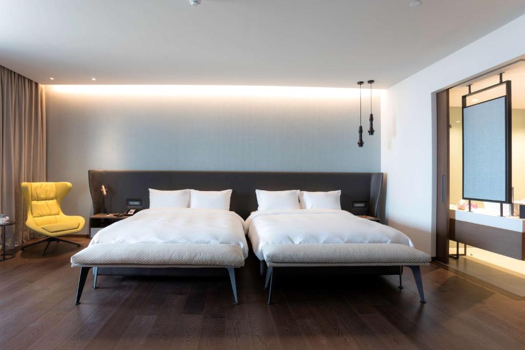 Mūsdienīgs viesnīcas numurs ar divām gultām, dzeltenu krēslu un elegantu apgaismojumu. Guļamistaba ir gaumīgi iekārtota ar koka grīdu un minimālu dekoru.