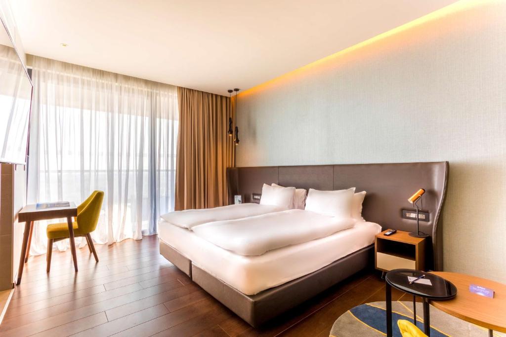 Mūsdienīga viesnīcas istaba ar divguļamo gultu, ērtu dīvānu un darba galdu, kas piedāvā plašu skatu caur logiem ar caurspīdīgām aizkariem.