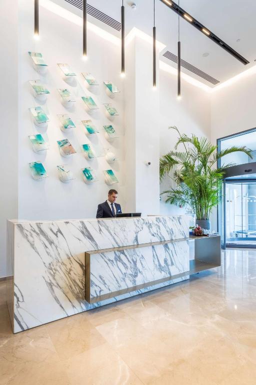 Mūsdienīgs viesnīcas reģistratūras interjers ar marmora letīti, dārza augiem un mākslinieciskām sienas dekorācijām. Viesnīcas darbinieks stāv pie letes, gatavs uzņemt viesus.