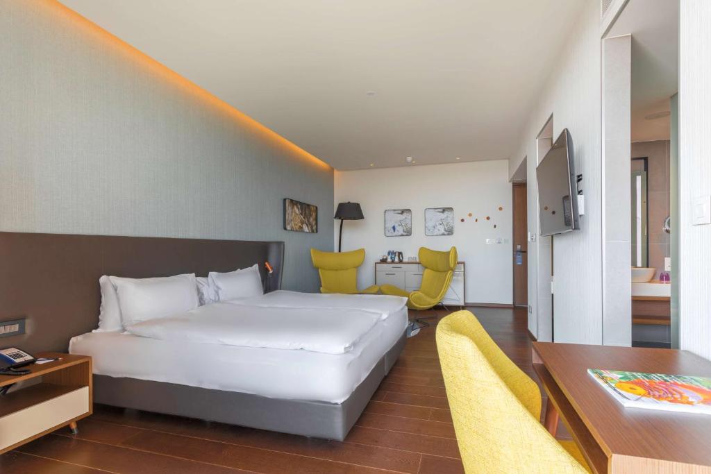 Mūsdienīgs viesnīcas numurs ar divguļamo gultu, dzeltenām krēslām un televizoru. Telpa ir gaiša un stilīga, radot komfortablu atmosfēru.