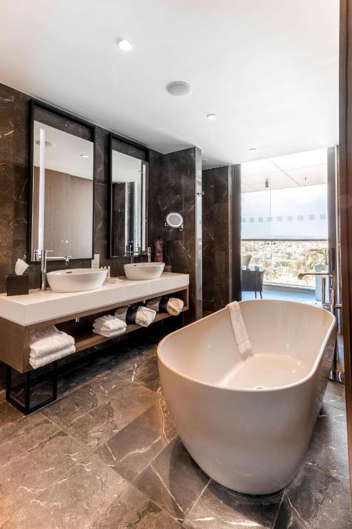 Mūsdienīgs vannas istabas dizains ar elegantu brīvo stūra vannu, divām izlietnēm un lieliem spoguļiem. Interjers ir veidots ar akmens flīzēm un koka akcentiem, radot greznu atmosfēru.