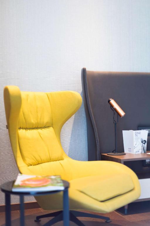 Dzeltena, mūsdienīga krēsla un melna dīvāna kompozīcija, blakus ir galdiņš ar žurnālu un lampu.