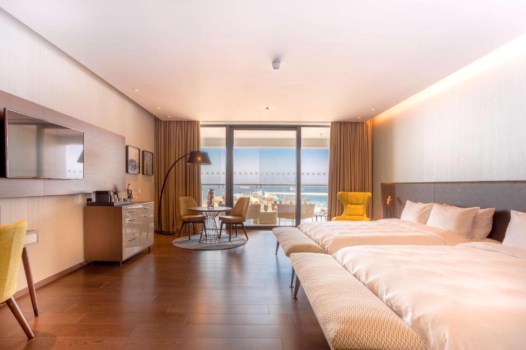 Mūsdienīga viesnīcas istaba ar divām gultām, dīvānu un ērtu krēslu, kas piedāvā skatu uz jūru. Interjers ir gaišs un elegants, ar koka grīdu un modernu apgaismojumu.