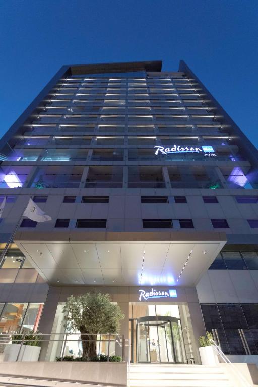 Moderns Radisson viesnīcas ēkas fasāde ar izgaismotu logo un ieeju, kas redzama vakara gaismā.