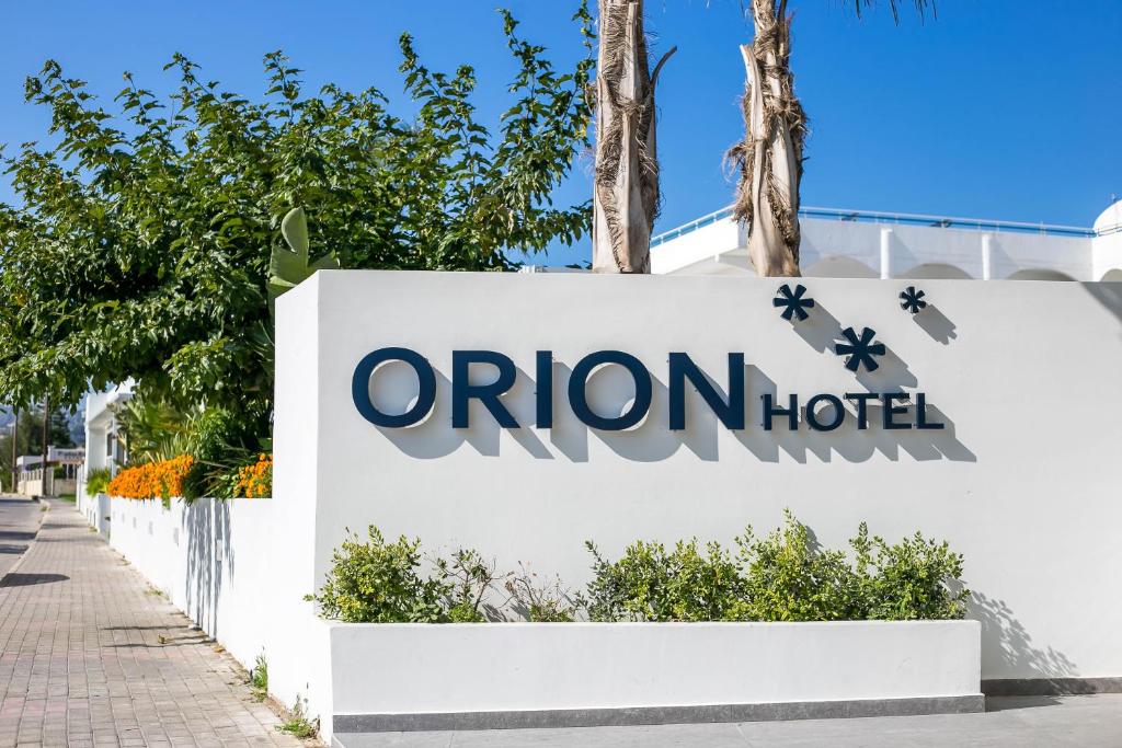 Orion Hotel zīme, kas atrodas pie viesnīcas ieejas, ieskauta zaļās augšanas un ziedu.