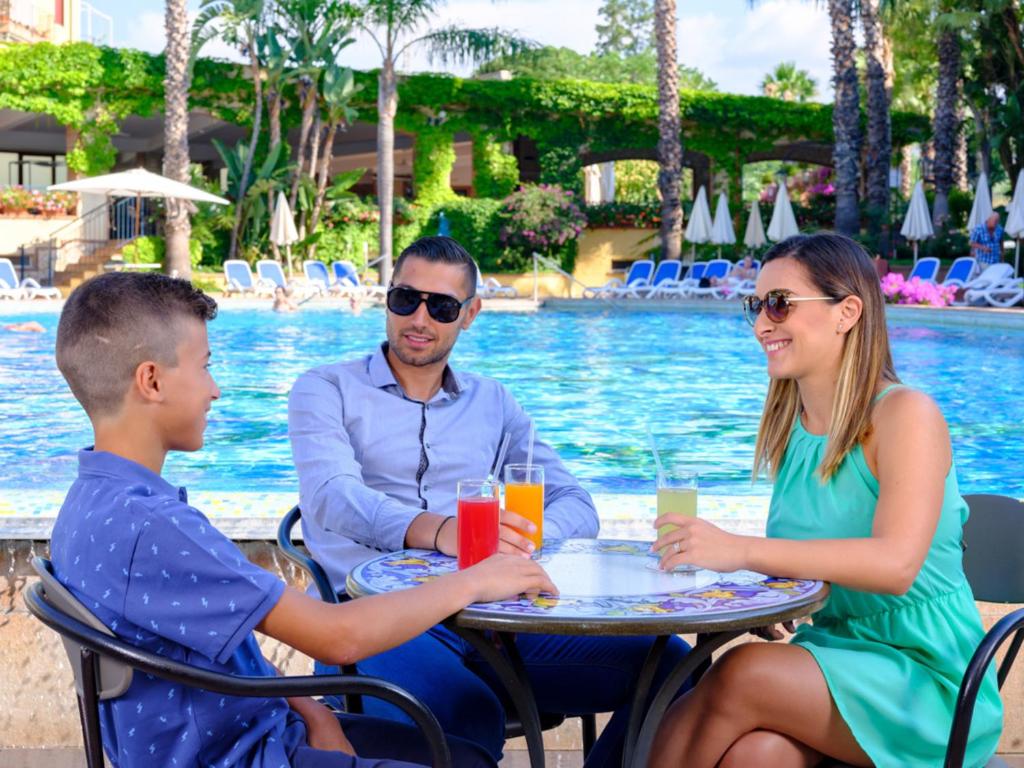 Trīs cilvēki sēž pie galda pie peldbaseina, baudot kokteiļus. Apkārt redzami palmām un saulainā atmosfērā.