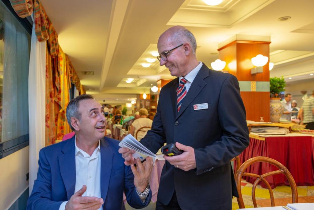 Vīrietis ar uzvalku un cits vīrietis ar viesmīļa uniformu sarunājas restorānā, izskatoties priecīgi un apmierināti. Apkārt redzami citi viesi un ēdienu galdi.