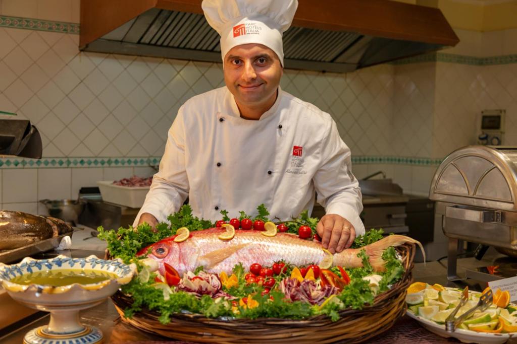 Šefpavārs pozē, turot rokās krāšņu zivju un dārzeņu kompozīciju, kas izkārtota uz liela trauka, kam blakus ir olīveļļa un citrons. Fonā redzama virtuve ar dažādiem pārtikas produktiem.