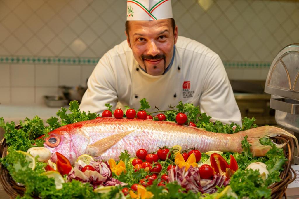 Šefpavārs pozē ar svaigu zivi, kas pasniegta uz dārzeņu gultas, ieskaitot tomātus un salātus. Fons ir virtuve, kas norāda uz kulinārijas prasmi.