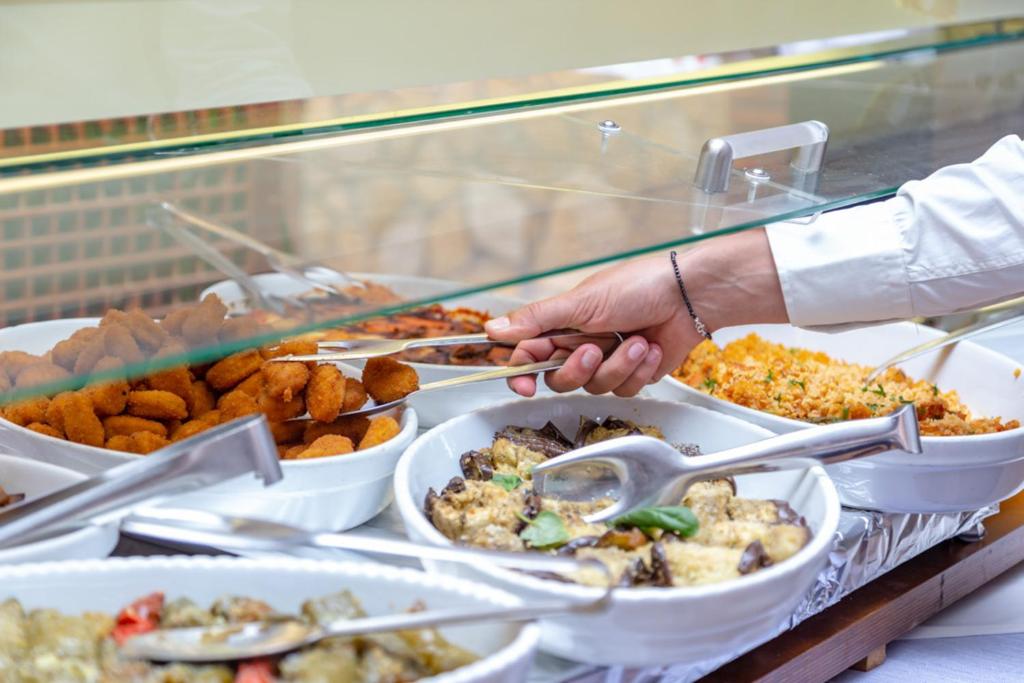 Svaiga ēdienu bufete ar dažādiem ēdieniem un cilvēka roku, kas ņem porciju ar tējkaroti.