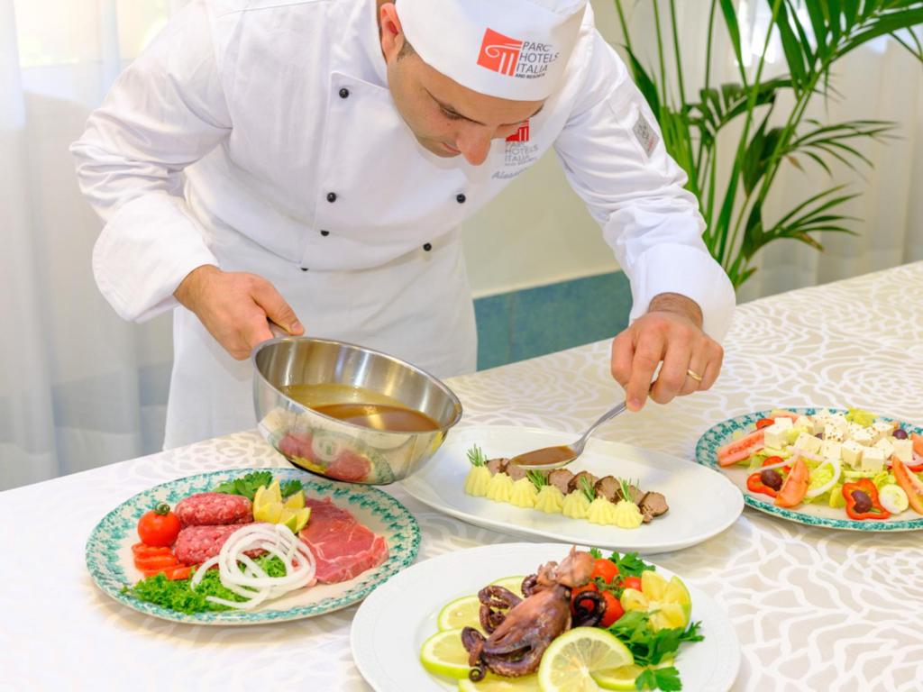 Šefpavārs gatavo izsmalcinātu ēdienu, pievienojot mērci uz šķīvja ar gaļu un kartupeļu biezeni, kam blakus ir dažādi uzkodu šķīvji ar jūras veltēm un dārzeņiem.