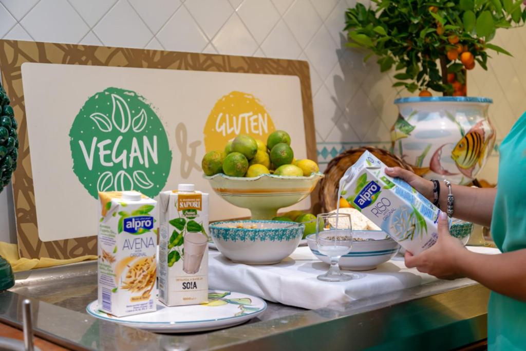 Veganisma un bezglutēna produktu prezentācija, kurā redzami dažādi augu izcelsmes dzērieni un augļi, kā arī krāsains dekors un trauki.