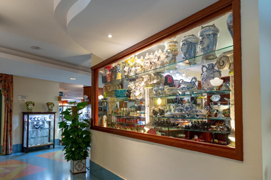 Stilīga vitrīna ar dažādiem mākslas priekšmetiem un keramikas izstrādājumiem, apgaismotā telpā ar krāsainu grīdu un dekoratīviem elementiem.