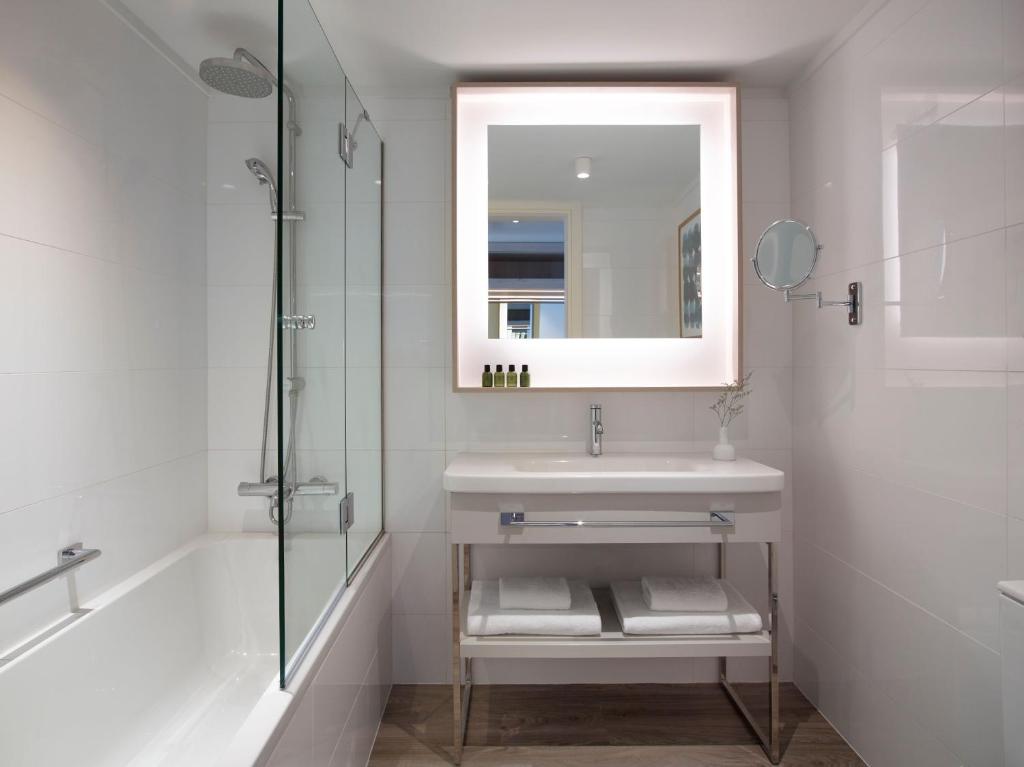 Mūsdienīgs vannas istabas interjers ar stikla dušu, vannu un modernu spoguli ar apgaismojumu. Uz izlietnes atrodas kosmētikas piederumi un tīri dvieļi.