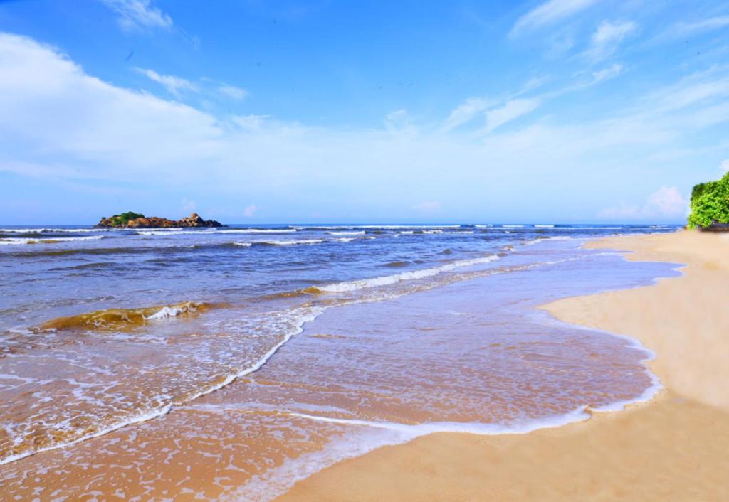 Smilšu pludmale ar mierīgu jūru un zilo debesu fona, kur viļņi maigā veidā sasniedz krastu. Tuvumā redzama maza klints sala, kas piešķir ainavai papildu šarmu.