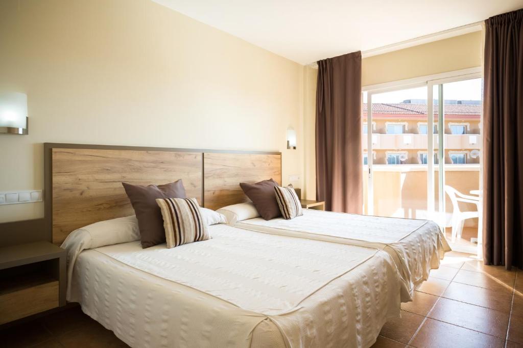 Mūsdienīga viesnīcas istaba ar divām gultām, ar koka galvām un dekoratīvajām spilveniem, kuru aizmugurē ir balkons ar skatu uz ārpusi. Gaismas toņi un vienkāršs dizains rada mājīgu atmosfēru.