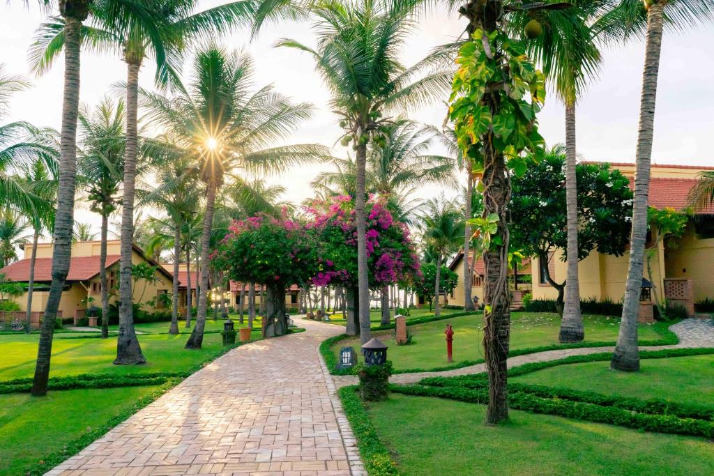 Tropu dārzs ar palmām un ziediem, kas ieskauj bruģētu ceļu, pie viesnīcas ar saules gaismu, kas spīd cauri lapotnei.