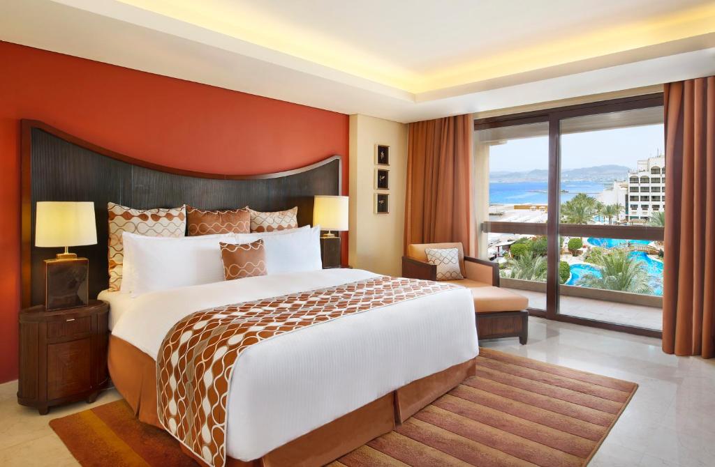 Mūsdienīga viesnīcas guļamistaba ar ērtu gultu un skatu uz baseinu un jūru, rotāta ar siltām krāsām un mīkstiem audumiem.