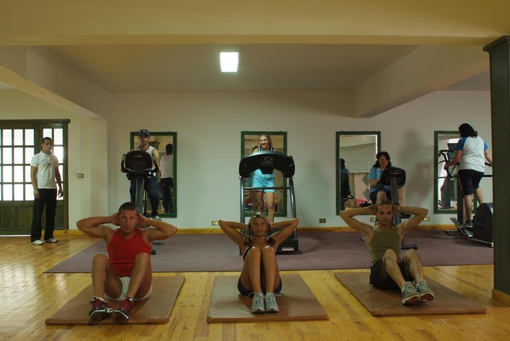 Sporta zāle ar cilvēkiem, kas nodarbojas ar vingrošanu uz paklājiem un izmanto trenažierus. Telpa ir gaiša, ar koka grīdu un spoguļiem sienās.