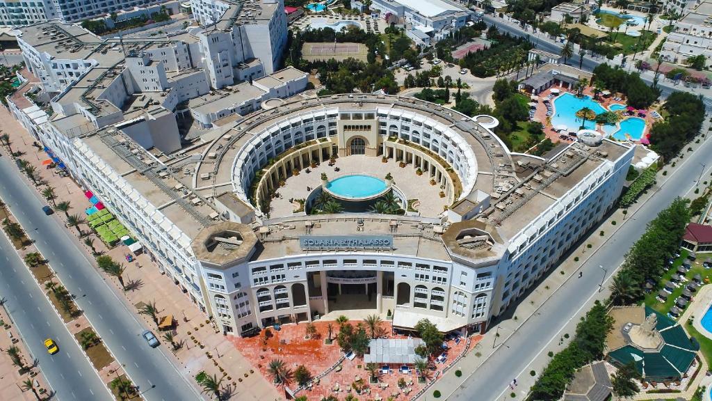Lielisks skats uz viesnīcu ar centrālo baseinu un apkārtējiem peldēšanas laukumiem. Ap ēku redzami zaļumi un ceļi ar palmām.