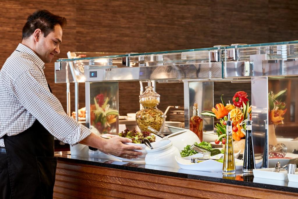 Vīrietis apkalpo ēdienus pie bufetes galda, kur redzami dažādi salāti un dārzeņi. Fonā ir krāšņas ziedu kompozīcijas un citi ēdieni.