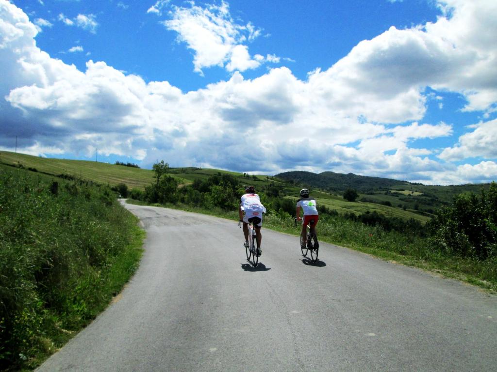 Divi riteņbraucēji brauc pa šauru ceļu, apņemti ar zaļojošu ainavu un zilo debesi ar mākoņiem.