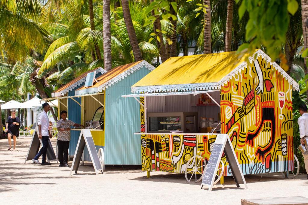 Krāsaini pārtikas stendi ar ainavu palmu fonā, kur cilvēki pasūta ēdienus. Šie stendi ir dekorēti ar dzīvīgu mākslu un sniedz ātrās uzkodas pludmales atmosfērā.