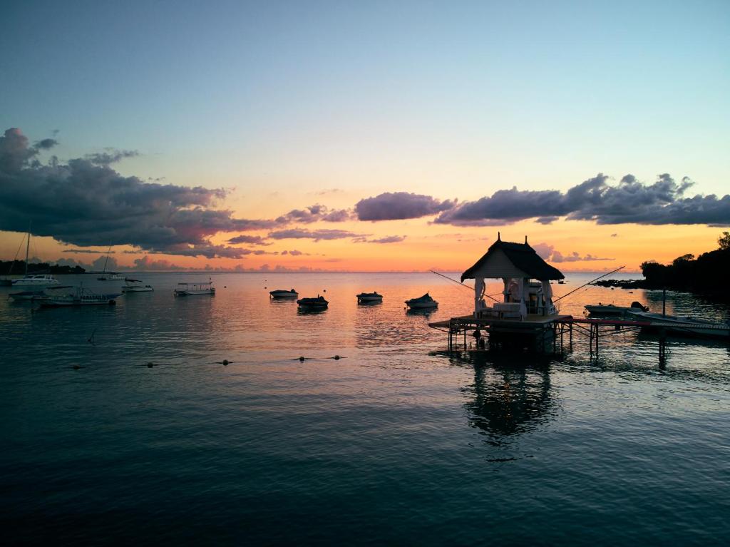 Saulrieta skats uz mierīgu jūru ar laivām un koka piestātni, kas atrodas pie krasta. Debesis ir krāsotas ar siltām oranžām un rozā nokrāsām.