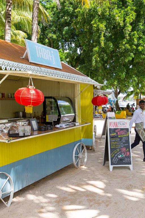 Krāsains ēdienu stends ar Āzijas ēdienu izvēli, kas izceļas ar sarkaniem laternām un izkārtnēm. Apkārt ir palmu koki, kas rada tropisku atmosfēru.
