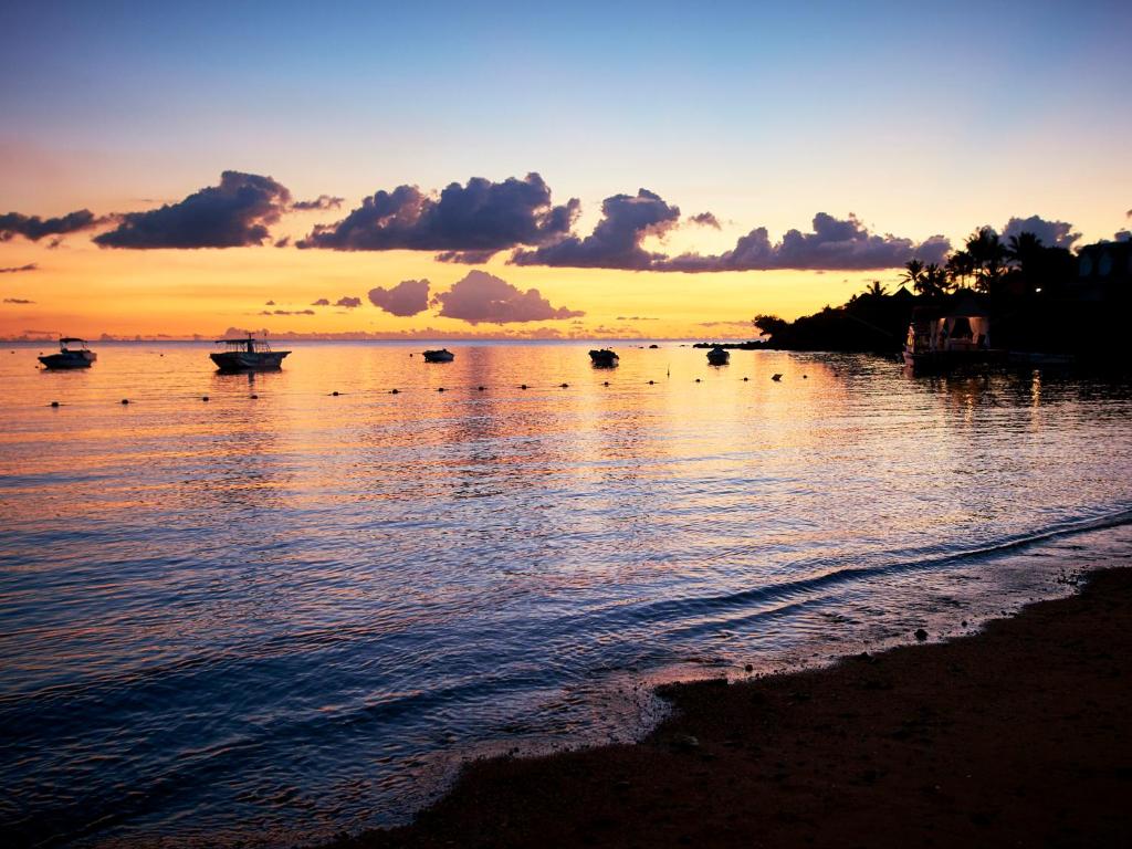Saulrieta skats pār mierīgu jūru ar laivām un siltiem toņiem. Kluss pludmales skats ar debesīm, kas iekrāsojas oranžā un zeltainā gaismā.