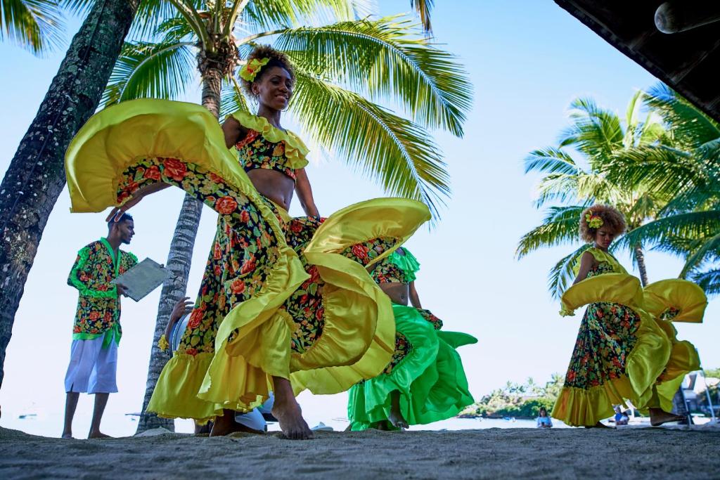 Deju priekšnesums tropiskā vidē ar krāsainām kleitām un palmām fonā. Tradicionālās dejas veido dzīvīgu atmosfēru pludmalē.