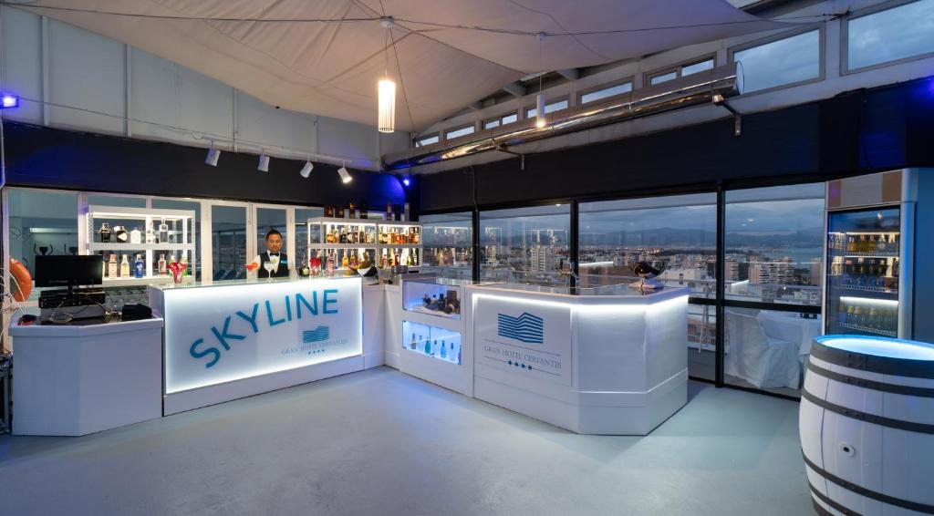 Elegantā bāra interjers ar panorāmas skatu, kurā redzams bārmenis un dažādu alkoholu izvēle uz plauktiem. Bārs atrodas augstākajā stāvā, piedāvājot viesiem unikālu pieredzi un relaksāciju.