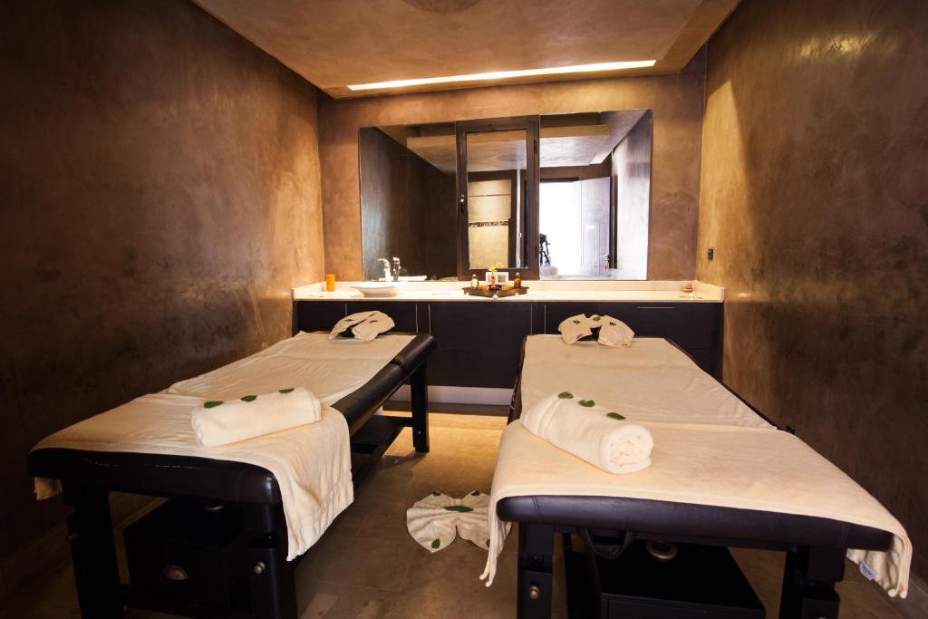 Relaksējoša masāžas telpa ar divām masāžas galdiem, kas rotāti ar dvieļiem un zaļām lapām, radot mierīgu atmosfēru. Interjers ir vienkāršs un moderns, ar tumšām sienām un elegantu izskatu.