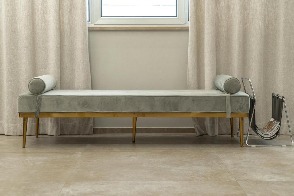 Mūsdienīgs dīvāns ar pelēku samta apdari un zeltainām kājām, novietots pie loga ar gaišām aizkariem. Blakus atrodas elegants žurnālu turētājs.