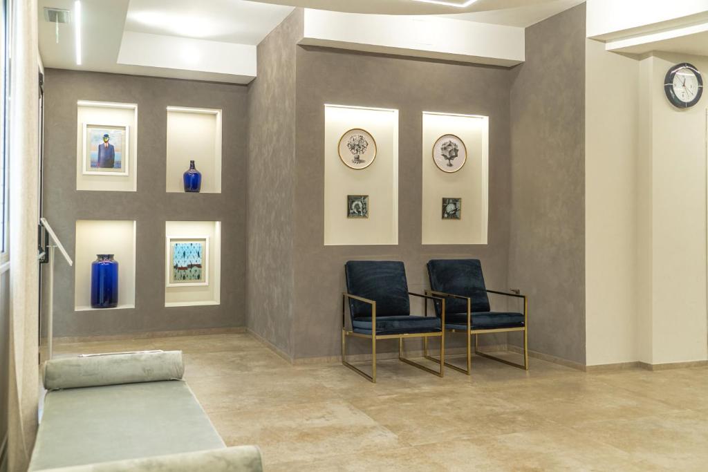 Mūsdienīgs interjers ar zilām dekoratīvām pudelēm un mākslas darbiem, kas izstādīti nišās uz pelēkas sienas. Divas dzeltenas krēsli gaida apmeklētājus.