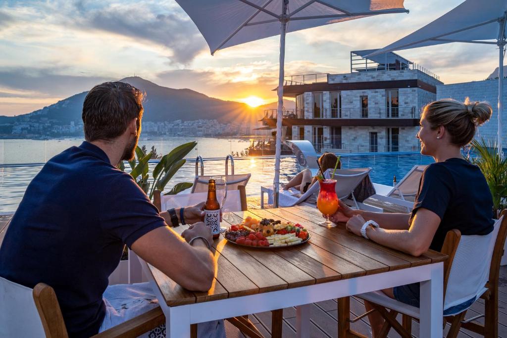 Romantiska saulrieta aina ar pāri sēdošiem cilvēkiem, kas bauda dzērienus un uzkodas pie baseina. Fons ar kalniem un mūsdienīgu arhitektūru papildina relaksējošo atmosfēru.