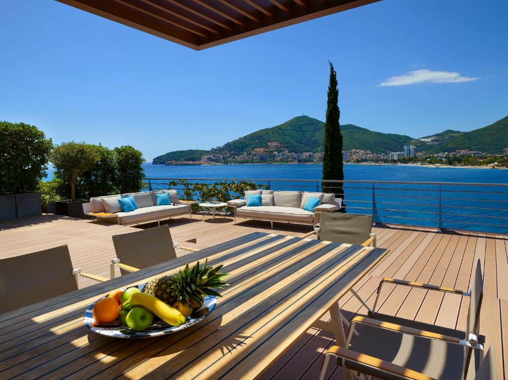 Lieliska terase ar skatu uz jūru, kurā ir moderni dīvāni un galda ēdamais ar augļiem. Fons veido kalnu ainava un saulainas debesis.