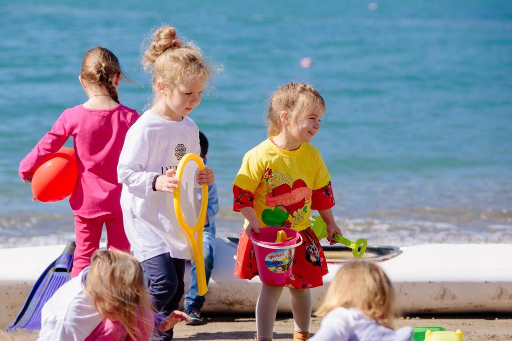 Bērni spēlējas pludmalē, izmantojot rotaļlietas, piemēram, smilšu spainīšus un riņķus, kamēr fons ir saulains un jūra.