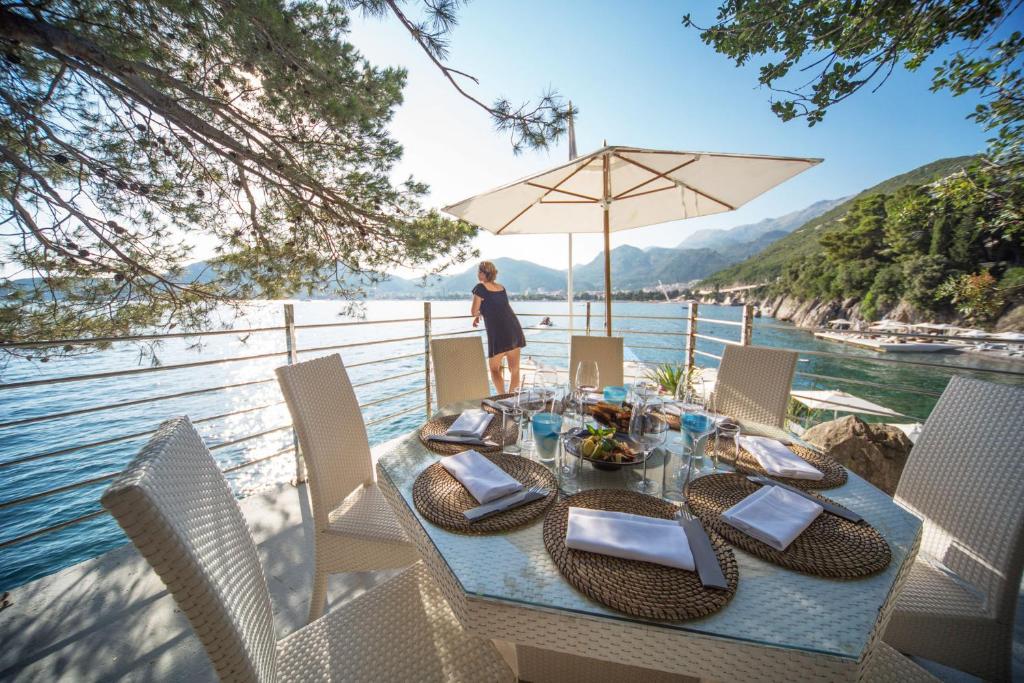 Romantiska ēdamzona pie jūras ar panorāmas skatu uz kalniem un ūdeni, kurā redzama sieviete, kas vēro dabu. Galdiņš ir klāts ar ēdieniem un dzērieniem, radot relaksējošu atmosfēru.