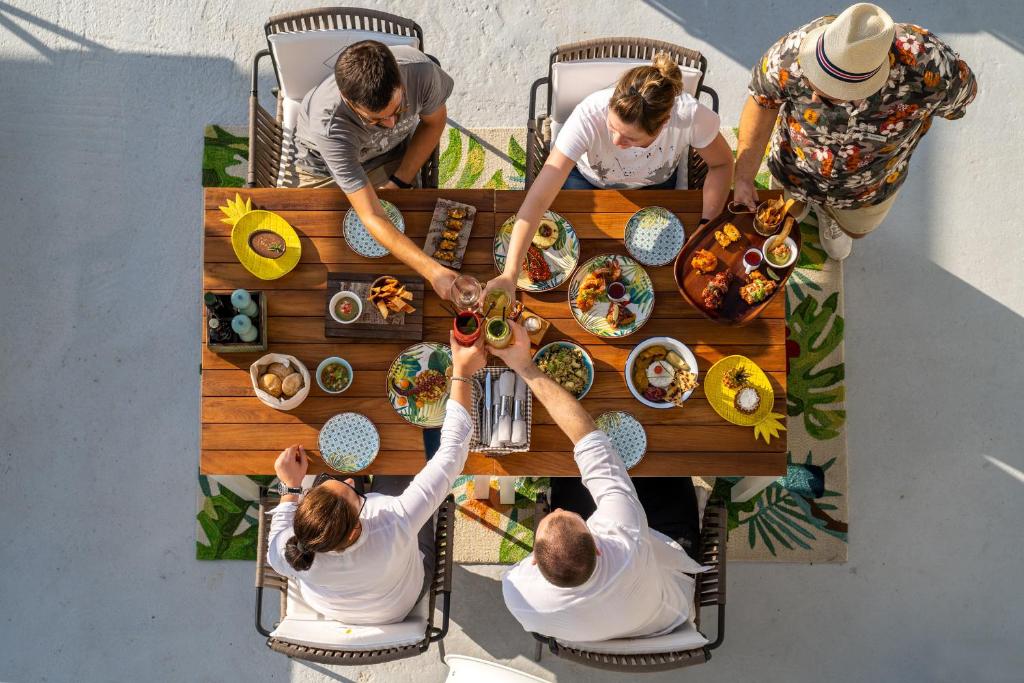 Pusdienu galds ar dažādiem ēdieniem un dzērieniem, ko dalās draugi saulainā vidē. Galds ir dekorēts ar krāsainām šķīvjiem un augļiem, radot svinīgu atmosfēru.