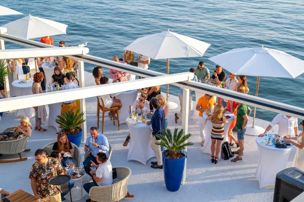 Vasaras svinības pie ūdens ar cilvēkiem, kas izbauda dzērienus un iepazīstas. Balti saulessargi un augi radījuši patīkamu atmosfēru.