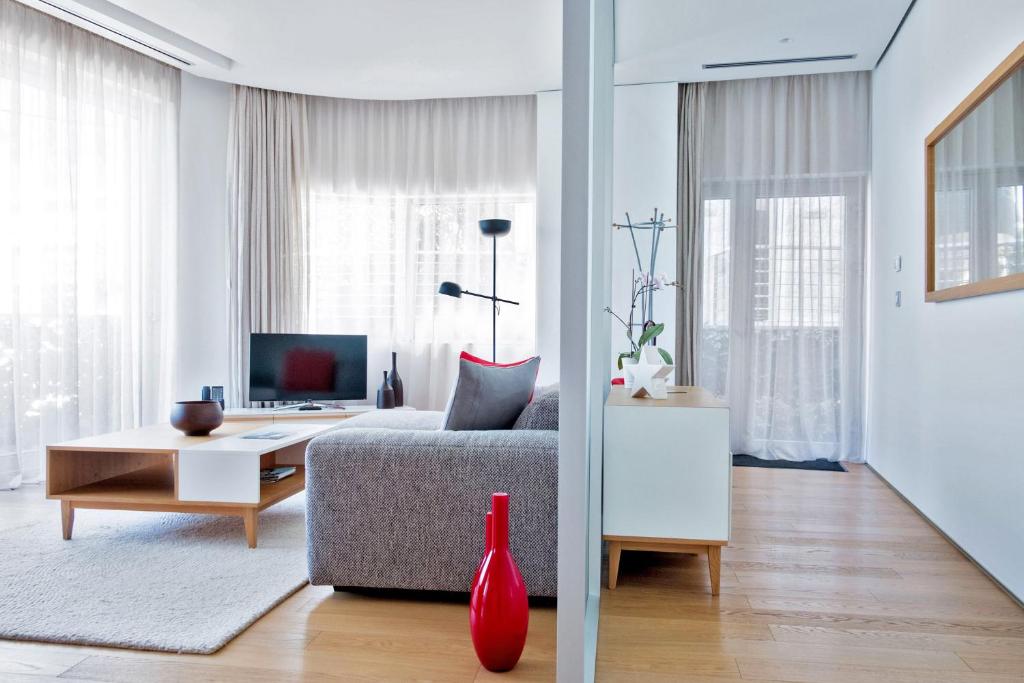 Mūsdienīgs viesistabas interjers ar pelēku dīvānu, koka kafijas galdiņu un televizoru. Telpa ir apgaismota ar dabīgu gaismu, un tajā ir redzami dekoratīvi elementi, piemēram, zieds un sarkana vāze.