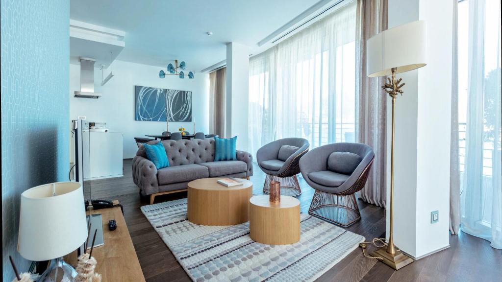 Mūsdienīgs viesistabas interjers ar dīvānu, divām krēslām un koka galdiņu, kas izceļs gaišajā un plašajā telpā ar lieliem logiem.