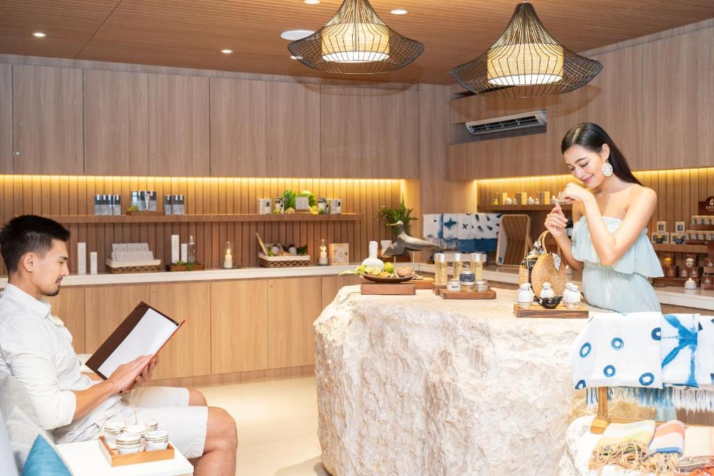 Mūsdienīga spa telpa ar diviem cilvēkiem, kur viens vīrietis izvēlas procedūras no saraksta, bet sieviete, tērpta eleganti, piedāvā produktus. Interjers ir gaišs ar koka apdari un dekoratīvām lampām.