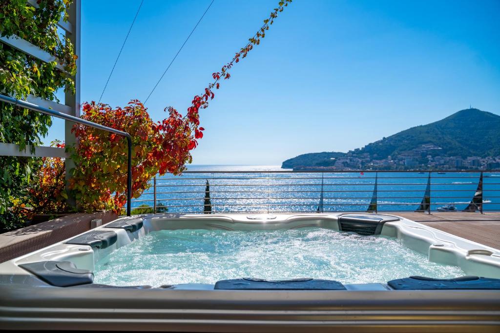 Apsildāms baseins ar burbuļiem, kas atrodas pie jūras, ar skaistu skatu uz kalnu un saulainu debesi. Apkārt aug krāšņas vīnogu lapas, radot relaksējošu atmosfēru.