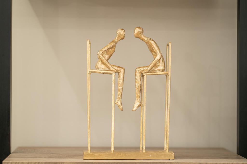 Zelta krāsas skulptūra, kurā attēloti divi cilvēki, kas sēž uz stabiem, simbolizējot tuvību un mijiedarbību.